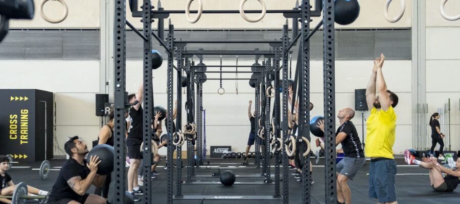 Estación rig en un ambiente de competición con personas entrenando y una serie de atletas realizando CrossFit Wall Ball.
